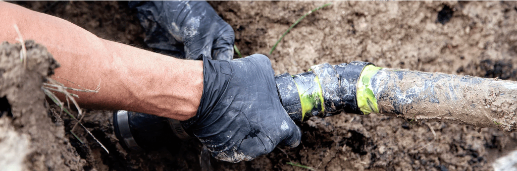 A plumber in Ottawa wearing black gloves works on repairing or connecting a muddy underground pipe wrapped with yellow tape.